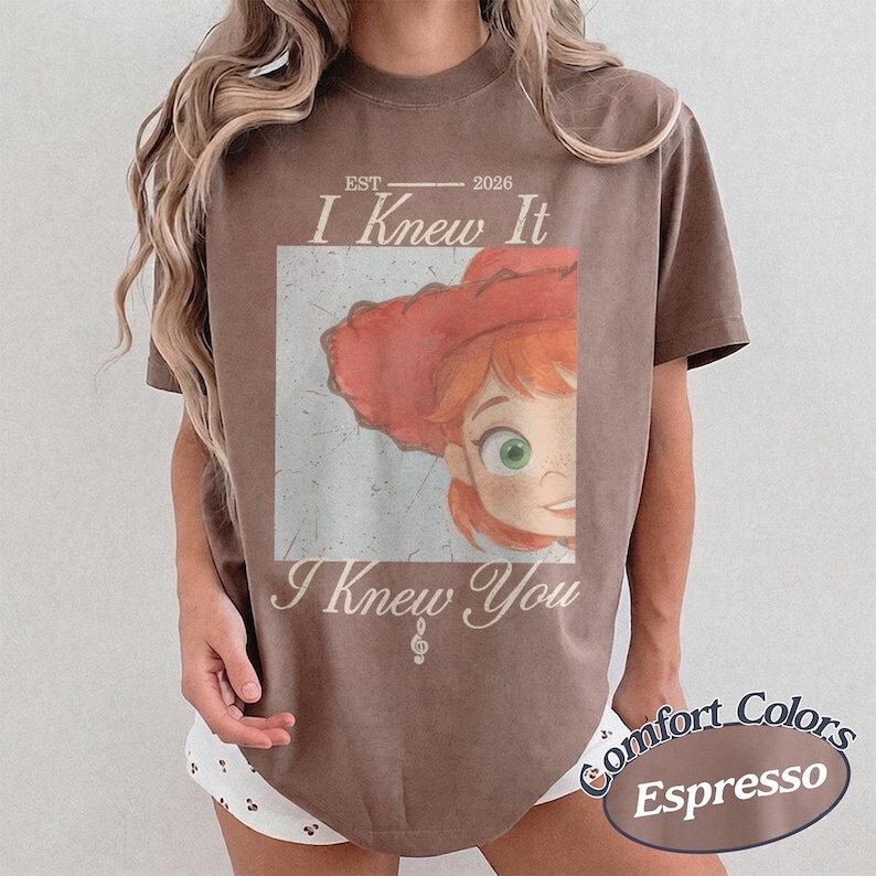 May include: A brown short-sleeve t-shirt featuring a graphic of a cartoon character with red hair and a red hat. The text reads EST 2026 I Knew It I Knew You with a small musical note symbol. The design has a distressed, vintage-style appearance.