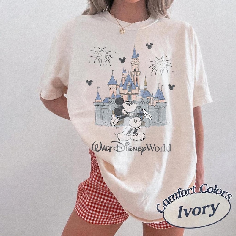 May include: Ivory-colored t-shirt featuring a graphic of Mickey Mouse in front of Cinderella's castle at Walt Disney World. The design includes fireworks and Mickey Mouse silhouettes. The shirt is labeled "Comfort Colors Ivory."
