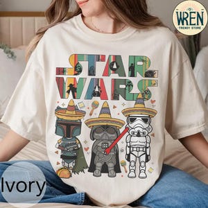 May include: A white t-shirt featuring a colourful "Star Wars" graphic. The graphic depicts the characters Boba Fett, Darth Vader, and a Stormtrooper wearing sombreros. The text "Star Wars" is in a colourful, retro font.