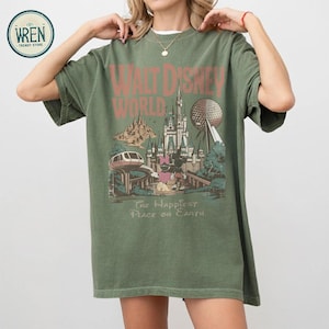 Może przedstawiać: Zielona koszulka w kolorze oliwkowym z grafiką w stylu vintage z Walt Disney World. Projekt zawiera nazwę parku, Zamek Kopciuszka, Spaceship Earth i frazę "The Happiest Place on Earth."