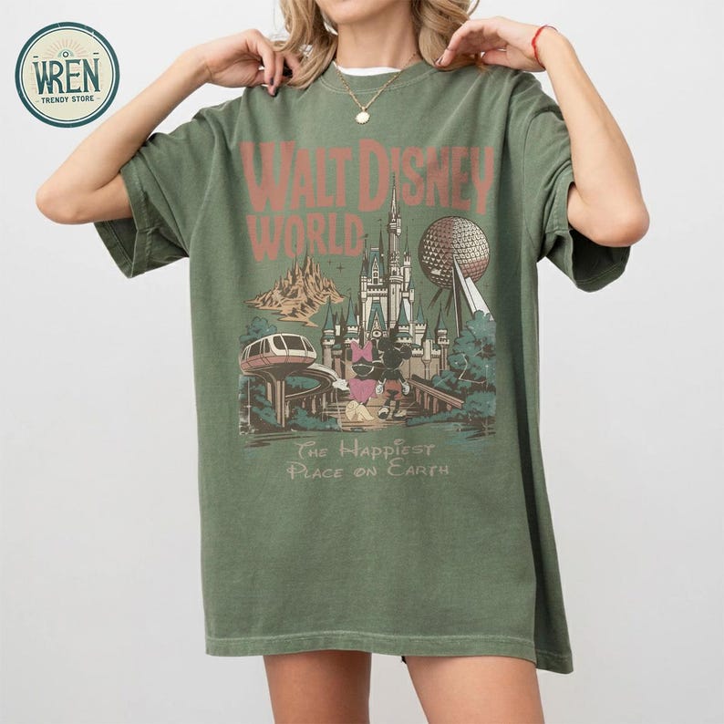 Może przedstawiać: Zielona koszulka w kolorze oliwkowym z grafiką w stylu vintage z Walt Disney World. Projekt zawiera nazwę parku, Zamek Kopciuszka, Spaceship Earth i frazę "The Happiest Place on Earth."