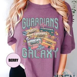 K&ouml;nnte beinhalten: Lilafarbenes T-Shirt mit einem grafischen Design aus Kassetten, Sternen, Planeten und einem UFO. Der Text "GUARDIANS of the GALAXY" ist auf dem Shirt aufgedruckt. Der Text auf den Kassetten lautet "hook on a feeling", "Come and get your love", "I want you back", "Spirit in the sky" und "I'd do anything for love".