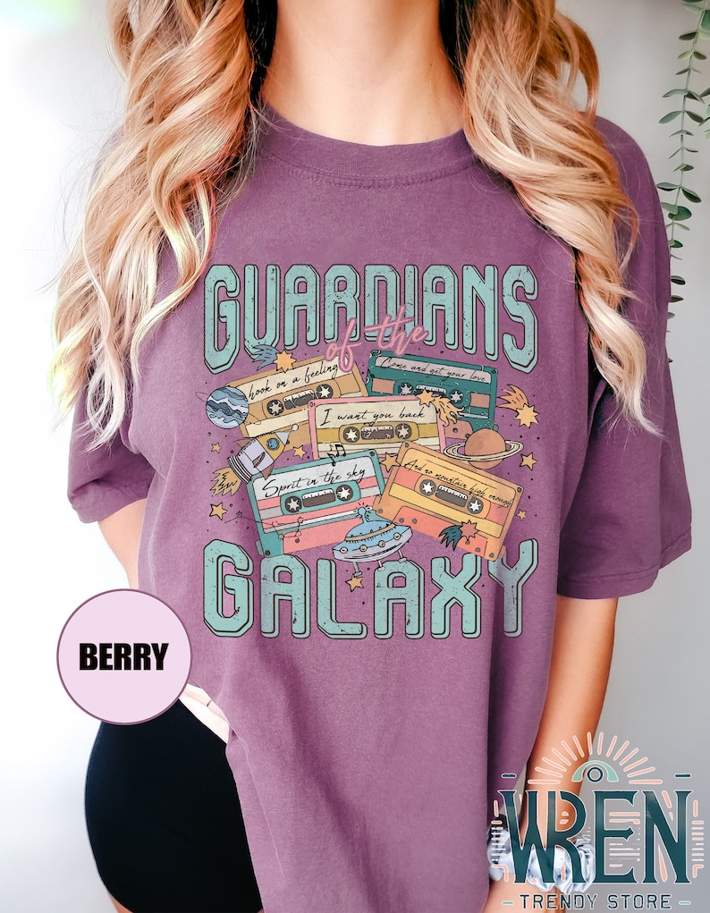 K&ouml;nnte beinhalten: Lilafarbenes T-Shirt mit einem grafischen Design aus Kassetten, Sternen, Planeten und einem UFO. Der Text "GUARDIANS of the GALAXY" ist auf dem Shirt aufgedruckt. Der Text auf den Kassetten lautet "hook on a feeling", "Come and get your love", "I want you back", "Spirit in the sky" und "I'd do anything for love".
