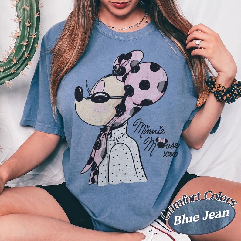 May include: Blue jean colored t-shirt featuring a Minnie Mouse graphic. The graphic shows Minnie Mouse wearing sunglasses and a polka dot bow and scarf. The text "Minnie Mouse xoxo" is below the graphic. The shirt has the words "Comfort Colors" and "Blue Jean" printed on it.