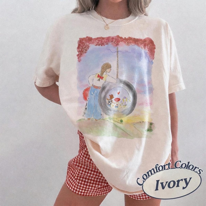 May include: An ivory short-sleeve t-shirt featuring a watercolor-style graphic of a person with braided hair standing next to a tire swing. The illustration includes a sunset sky, red foliage, and small figures inside the tire. The shirt has a crew neck.