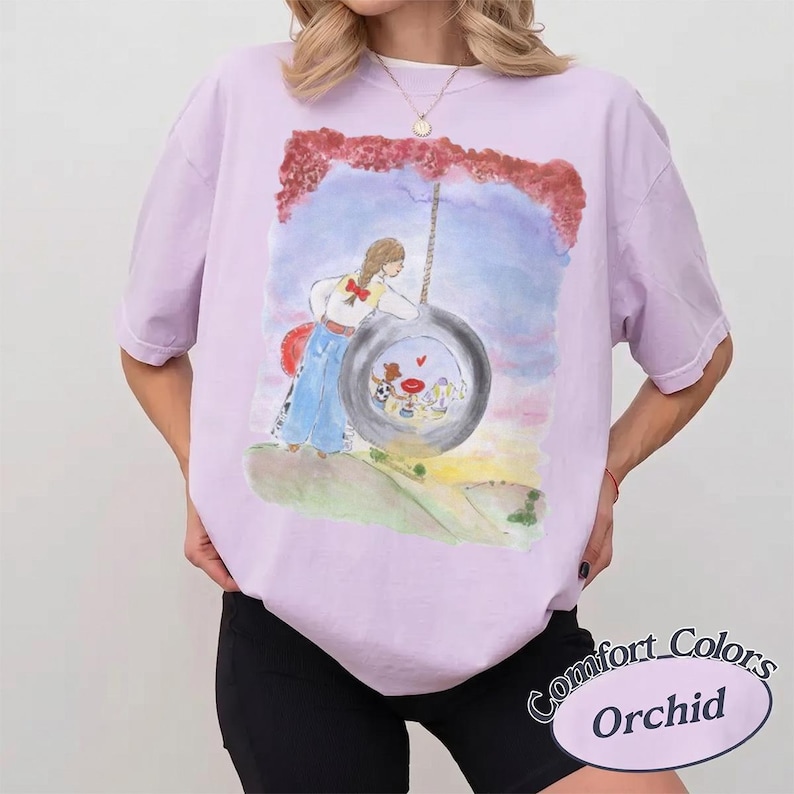 May include: An orchid-colored t-shirt featuring a watercolor-style graphic of a person standing next to a tire swing. The illustration includes a blue sky, a grassy hill, and a small red heart symbol. The text Comfort Colors Orchid is visible.