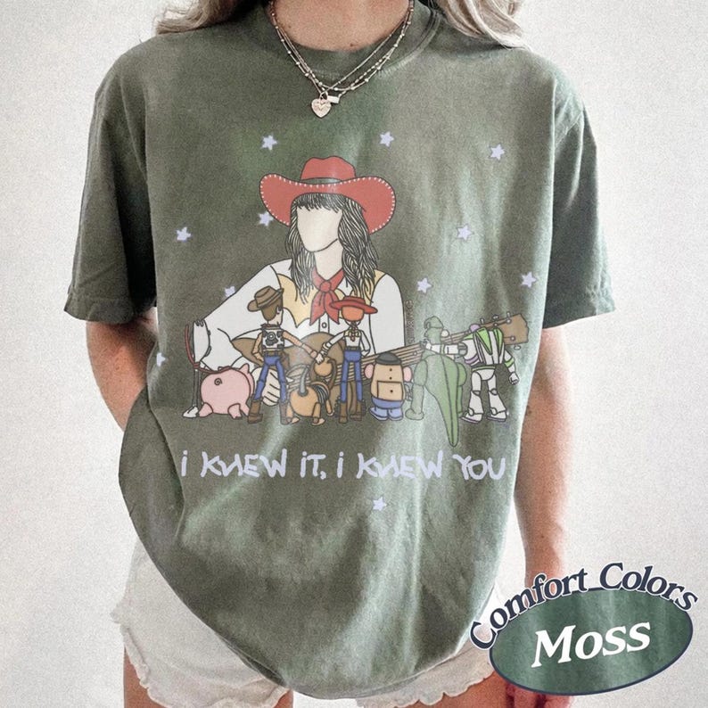 May include: An olive green t-shirt featuring a graphic illustration of a person in a red cowboy hat surrounded by various toy characters, including a pig, a space ranger, and a dinosaur. The text "I KNEW IT, I KNEW YOU" is printed below the graphic.