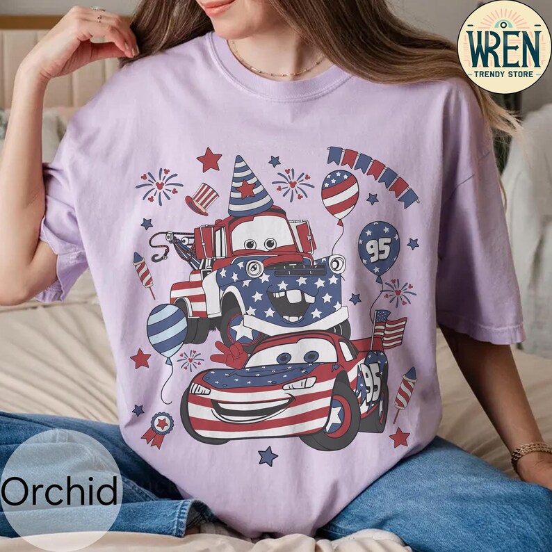 Puede incluir: Camiseta lila claro con un dise&ntilde;o patri&oacute;tico con dos coches de dibujos animados, estrellas, fuegos artificiales y globos. Los coches est&aacute;n decorados con patrones rojos, blancos y azules, y el n&uacute;mero "95" es visible.