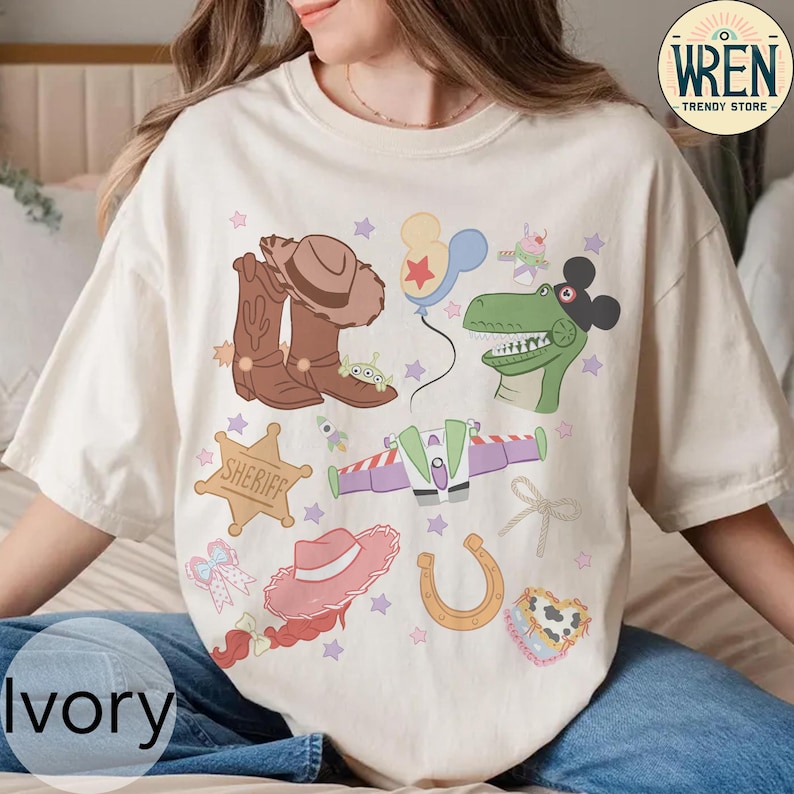 May include: Ivory-colored t-shirt featuring a collection of cartoon illustrations. The design includes a cowboy hat, boots, a sheriff badge, a dinosaur, a spaceship, and other playful elements. The text "WREN TRENDY STORE" is in the upper right corner.