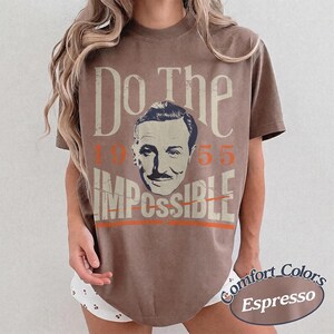 May include: Espresso-colored t-shirt with the text "Do The Impossible" and a portrait of a man. The numbers "1955" are in orange. The shirt is part of the Comfort Colors collection.