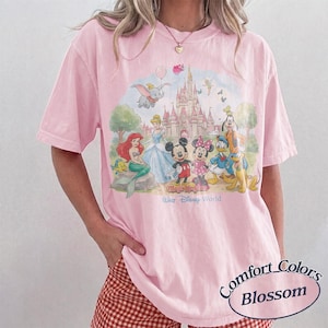 May include: A light pink short-sleeve t-shirt featuring a colorful graphic of Disney characters including Mickey Mouse, Minnie Mouse, Ariel, Cinderella, Goofy, Donald Duck, and Dumbo in front of a castle with the text Magic Kingdom Walt Disney World.