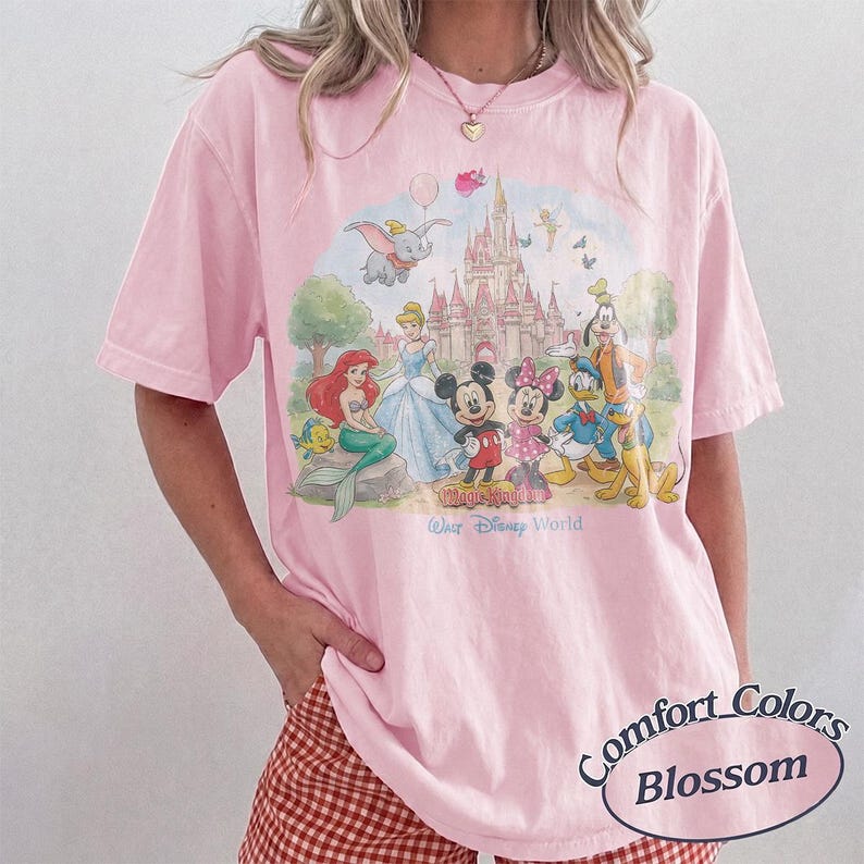 May include: A light pink short-sleeve t-shirt featuring a colorful graphic of Disney characters including Mickey Mouse, Minnie Mouse, Ariel, Cinderella, Goofy, Donald Duck, and Dumbo in front of a castle with the text Magic Kingdom Walt Disney World.