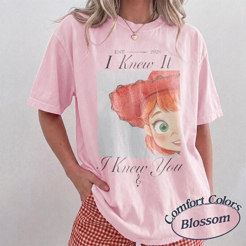May include: A light pink short-sleeve t-shirt featuring a graphic print of a cartoon character with red hair and a red hat. The text reads EST 2026, I Knew It, and I Knew You. The shirt has a crew neckline and a relaxed fit, showcasing a casual style.