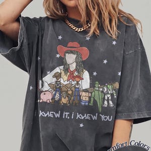 May include: A charcoal grey short-sleeve t-shirt featuring a graphic of a person in a cowboy hat playing a guitar surrounded by animated toy characters. The text "I KNEW IT, I KNEW YOU" is printed below the graphic, with "Comfort Colors Pepper" text.