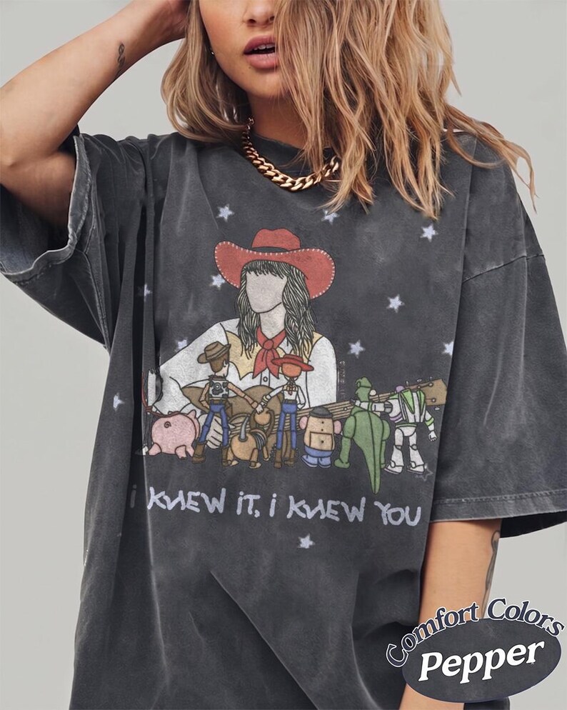 May include: A charcoal grey short-sleeve t-shirt featuring a graphic of a person in a cowboy hat playing a guitar surrounded by animated toy characters. The text "I KNEW IT, I KNEW YOU" is printed below the graphic, with "Comfort Colors Pepper" text.