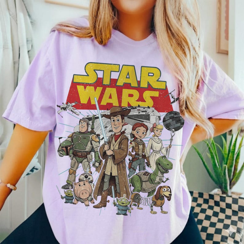 May include: A light purple t-shirt featuring a graphic of Star Wars characters and the Star Wars logo in yellow and red. The graphic includes characters from Toy Story and Star Wars.