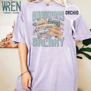 K&ouml;nnte beinhalten: Ein lavendelfarbenes T-Shirt mit einem Grafikdruck von Kassetten und dem Text "Guardians of the Galaxy". Die Grafik enth&auml;lt auch Planeten, Sterne und ein Raumschiff.