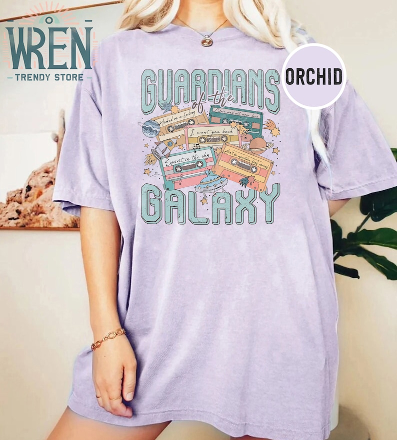 K&ouml;nnte beinhalten: Ein lavendelfarbenes T-Shirt mit einem Grafikdruck von Kassetten und dem Text "Guardians of the Galaxy". Die Grafik enth&auml;lt auch Planeten, Sterne und ein Raumschiff.