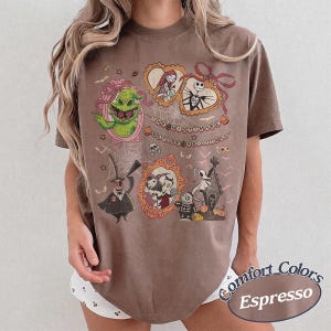 May include: Espresso-colored t-shirt featuring a graphic design from "The Nightmare Before Christmas." The design includes characters like Jack Skellington, Sally, and Oogie Boogie in ornate frames, with the film's title displayed above. The shirt is a Comfort Colors brand.