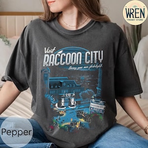Könnte beinhalten: Ein dunkelgraues T-Shirt mit einem Grafikdruck des Raccoon City Police Department Gebäudes aus der Resident Evil Videospielserie. Der Text "Visit Raccoon City" und "Bring your own flashlight" ist auf dem Shirt gedruckt.