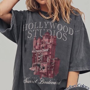 May include: A charcoal gray short-sleeve t-shirt featuring a graphic of the Hollywood Tower Hotel. The text reads HOLLYWOOD STUDIOS, The HOLLYWOOD TOWER, and AMID THE GLITZ AND GLAM Sunset Boulevard. The bottom corner includes the text Comfort Colors Pepper.