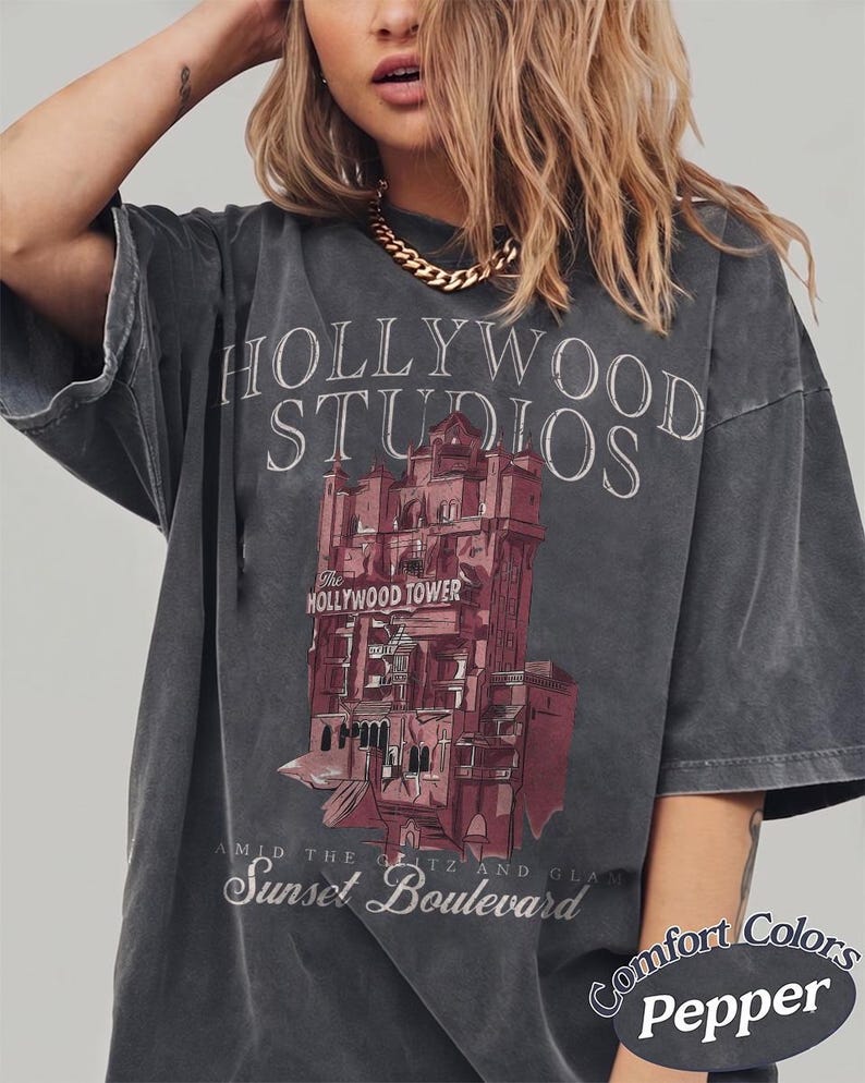 May include: A charcoal gray short-sleeve t-shirt featuring a graphic of the Hollywood Tower Hotel. The text reads HOLLYWOOD STUDIOS, The HOLLYWOOD TOWER, and AMID THE GLITZ AND GLAM Sunset Boulevard. The bottom corner includes the text Comfort Colors Pepper.