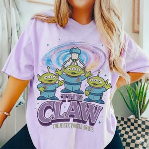 以下が含まれることがあります： 紫色のTシャツ。3つの目を持つ緑色の宇宙人が3人、青い制服を着て、クレーンゲーム機を持っています。「THE CLAW」の文字が下にあり、その下に「THE MYSTIC PORTAL AWAITS」と書かれています。背景にはカラフルな銀河のデザインがあります。