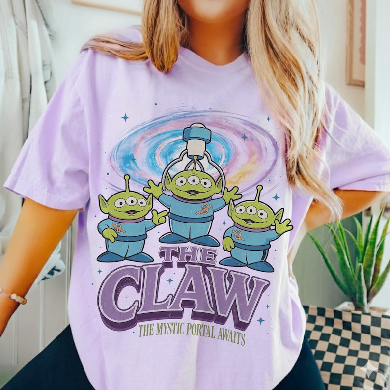 以下が含まれることがあります： 紫色のTシャツ。3つの目を持つ緑色の宇宙人が3人、青い制服を着て、クレーンゲーム機を持っています。「THE CLAW」の文字が下にあり、その下に「THE MYSTIC PORTAL AWAITS」と書かれています。背景にはカラフルな銀河のデザインがあります。