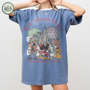 以下が含まれることがあります： ディズニーワールドのキャラクターとランドマークのグラフィックが描かれた青いTシャツ。デザインにはミッキーマウスとミニーマウス、ドナルドダックとグーフィーが含まれており、「Walt Disney World」と「The happiest place on earth」の文字が書かれています。