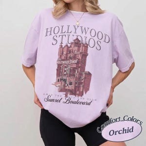May include: A light purple short-sleeve t-shirt featuring a dark graphic of the Hollywood Tower Hotel. The text reads Hollywood Studios, The Hollywood Tower, and Amid the Glitz and Glam Sunset Boulevard. The shirt is paired with black shorts.