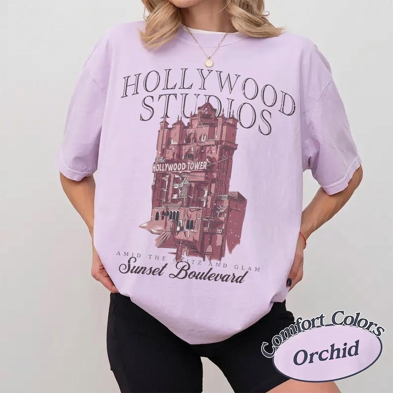 May include: A light purple short-sleeve t-shirt featuring a dark graphic of the Hollywood Tower Hotel. The text reads Hollywood Studios, The Hollywood Tower, and Amid the Glitz and Glam Sunset Boulevard. The shirt is paired with black shorts.