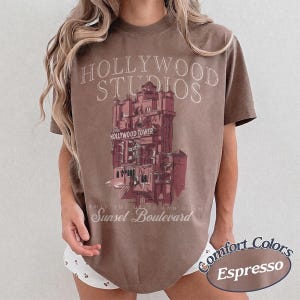 May include: A brown short-sleeve t-shirt featuring a graphic of the Hollywood Tower Hotel. The text reads Hollywood Studios, The Hollywood Tower, and Sunset Boulevard. The shirt has a crew neckline and a relaxed fit, with a vintage-inspired design.
