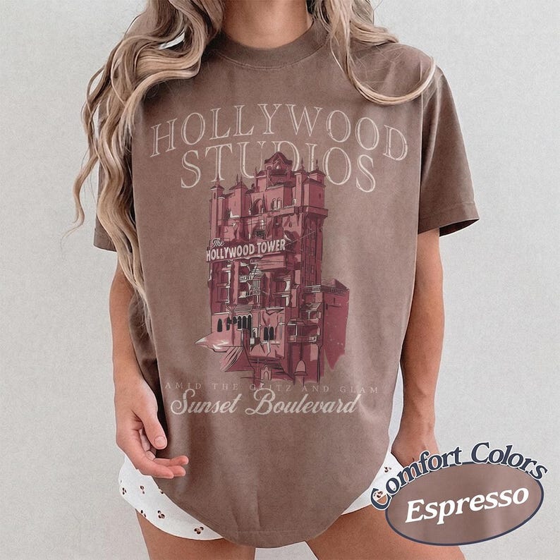 May include: A brown short-sleeve t-shirt featuring a graphic of the Hollywood Tower Hotel. The text reads Hollywood Studios, The Hollywood Tower, and Sunset Boulevard. The shirt has a crew neckline and a relaxed fit, with a vintage-inspired design.