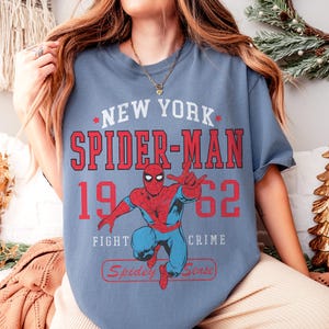 Op de afbeelding: Blauw T-shirt met een Spider-Man-afbeelding. Het ontwerp bevat de tekst "NEW YORK SPIDER-MAN 1962" in rood en wit, met de zinnen "FIGHT CRIME" en "Spidey Sense". De superheld is in een dynamische pose afgebeeld.
