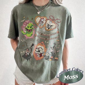 May include: A moss green t-shirt featuring characters from "The Nightmare Before Christmas." The design includes framed portraits of characters like Jack Skellington, Sally, and Oogie Boogie, with the film's title in a circular arrangement.