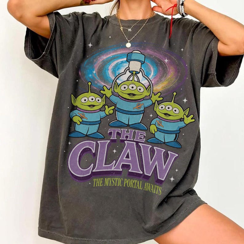 以下が含まれることがあります： 青い衣装を着た3人の緑色のエイリアンのグラフィックと「THE CLAW」の文字、その下に「THE MYSTIC PORTAL AWAITS」が描かれた濃い灰色のTシャツ。背景にはカラフルな銀河のデザインが描かれています。