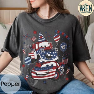 Puede incluir: Camiseta gris oscuro con un dise&ntilde;o patri&oacute;tico de dos coches de dibujos animados decorados con banderas y estrellas americanas. El dise&ntilde;o incluye globos, fuegos artificiales y el n&uacute;mero 95. La camiseta tiene la palabra "Pepper" impresa en la parte inferior izquierda.