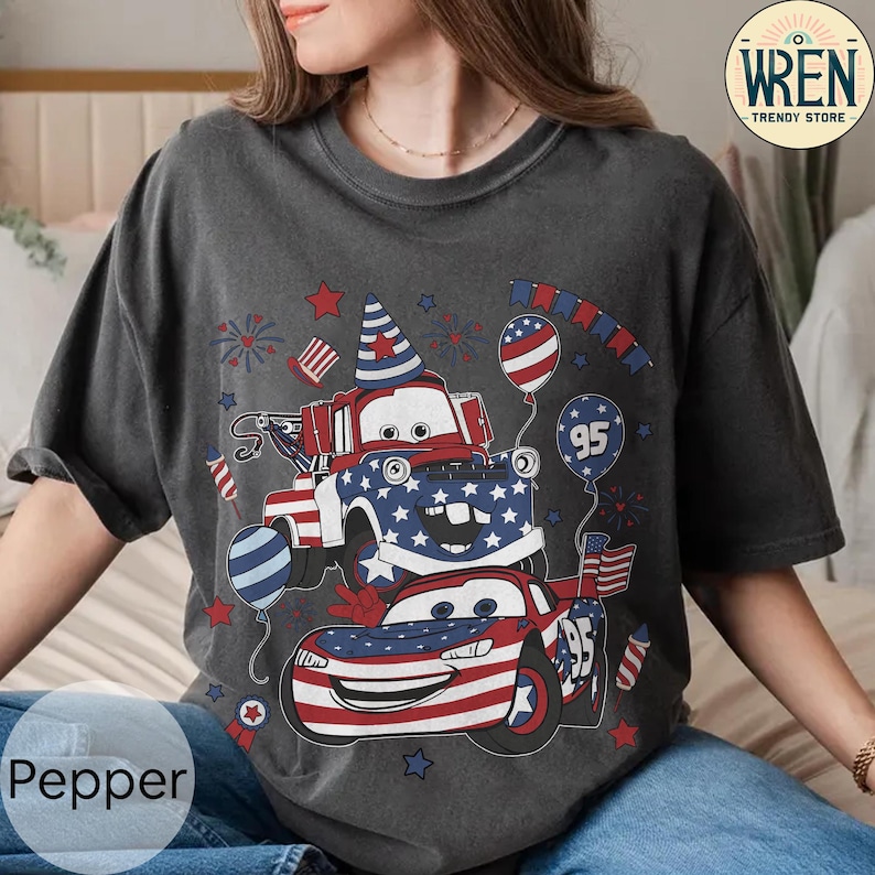 Puede incluir: Camiseta gris oscuro con un dise&ntilde;o patri&oacute;tico de dos coches de dibujos animados decorados con banderas y estrellas americanas. El dise&ntilde;o incluye globos, fuegos artificiales y el n&uacute;mero 95. La camiseta tiene la palabra "Pepper" impresa en la parte inferior izquierda.