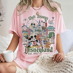 May include: Pink t-shirt featuring a colorful Disneyland graphic. The design includes Mickey Mouse, Minnie Mouse, Donald Duck, and other Disney characters in front of a Ferris wheel and castle. The text "Walt Disney's Disneyland Est. 1955" is also visible.