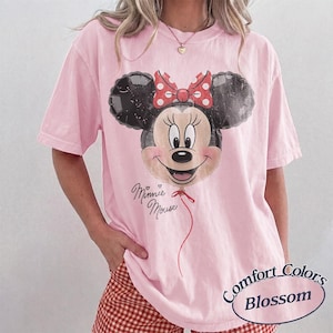 Op de afbeelding: Zachtroze Comfort Colors Blossom T-shirt met een Minnie Mouse ballon ontwerp. Het ontwerp bevat een rode strik, een hart en de woorden "Minnie Mouse". Het shirt heeft korte mouwen en een losse pasvorm.