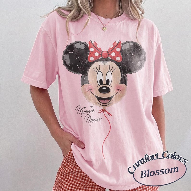 Op de afbeelding: Zachtroze Comfort Colors Blossom T-shirt met een Minnie Mouse ballon ontwerp. Het ontwerp bevat een rode strik, een hart en de woorden "Minnie Mouse". Het shirt heeft korte mouwen en een losse pasvorm.