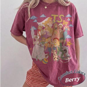 May include: A berry-colored short-sleeve t-shirt featuring a large graphic print of various animated characters in a collage style. The design includes the text TS, a rocket, a yellow star, and a colorful ball. The shirt has a crew neckline.