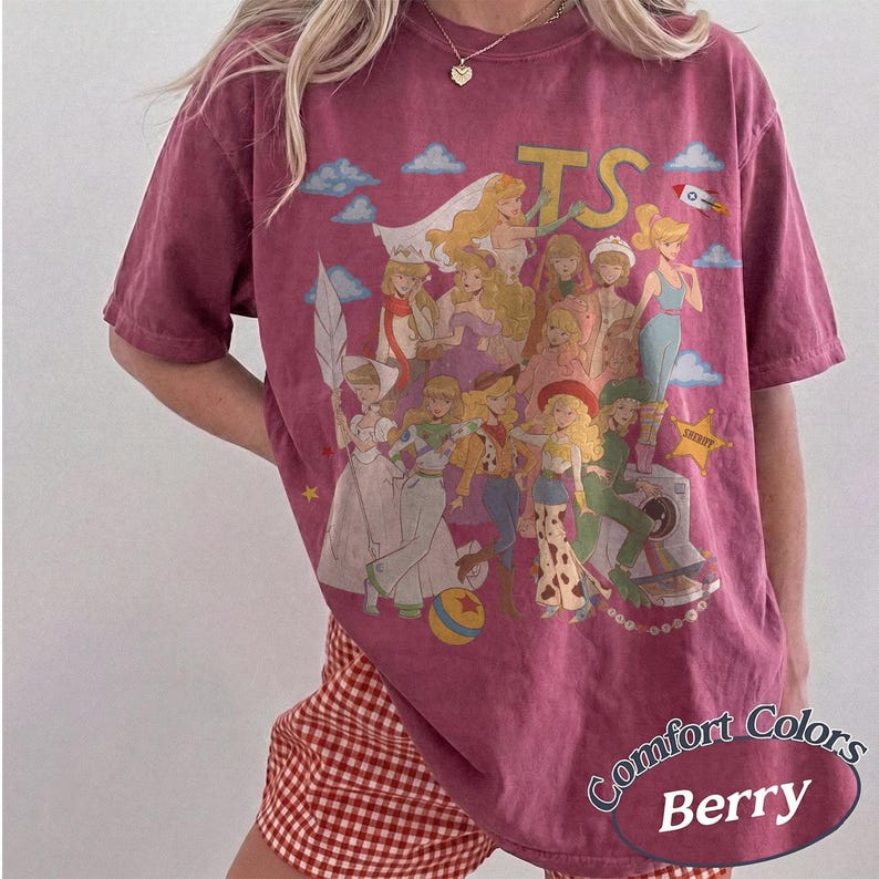May include: A berry-colored short-sleeve t-shirt featuring a large graphic print of various animated characters in a collage style. The design includes the text TS, a rocket, a yellow star, and a colorful ball. The shirt has a crew neckline.