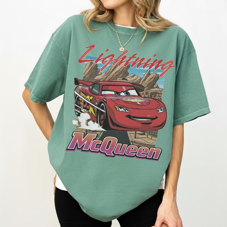 May include: Green t-shirt featuring a graphic of Lightning McQueen from the movie Cars. The design includes the text "Lightning McQueen" in red and purple, with a cartoon image of the red race car. The shirt has short sleeves and a relaxed fit.