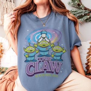 以下が含まれることがあります： 青いTシャツ。3つの目を持つ3人の緑色のエイリアンのグラフィック、青い制服を着て、クレーンゲームがあります。「THE CLAW」の文字は紫で表示され、その下に「THE MYSTIC PORTAL AWAITS」と書かれています。