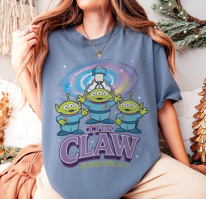 以下が含まれることがあります： 青いTシャツ。3つの目を持つ3人の緑色のエイリアンのグラフィック、青い制服を着て、クレーンゲームがあります。「THE CLAW」の文字は紫で表示され、その下に「THE MYSTIC PORTAL AWAITS」と書かれています。