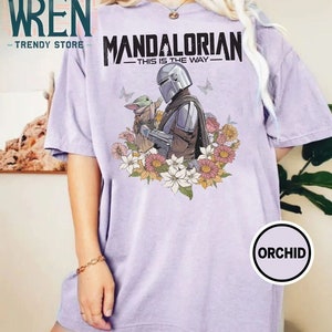 May include: A lavender t-shirt featuring a graphic of the Mandalorian and Grogu, surrounded by flowers. The text "Mandalorian - This is the Way -" is printed on the shirt.