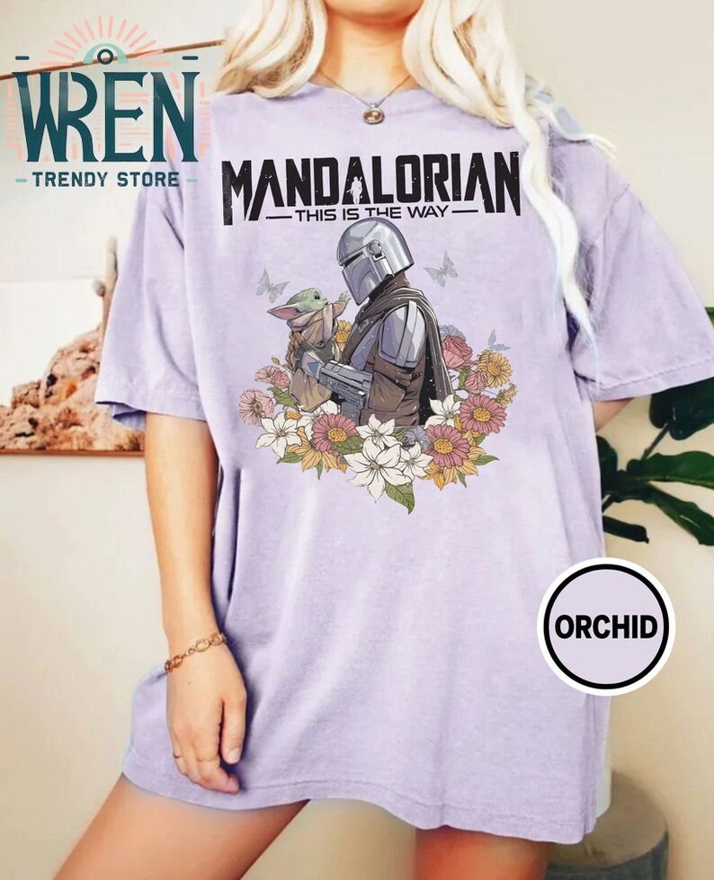 May include: A lavender t-shirt featuring a graphic of the Mandalorian and Grogu, surrounded by flowers. The text "Mandalorian - This is the Way -" is printed on the shirt.