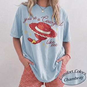 May include: A light blue t-shirt featuring a graphic of a red cowgirl hat with a white band and a red braided ponytail. The text reads you're a Cowgirl like me in a cursive red font, accented by small yellow stars. The shirt has a distressed print.