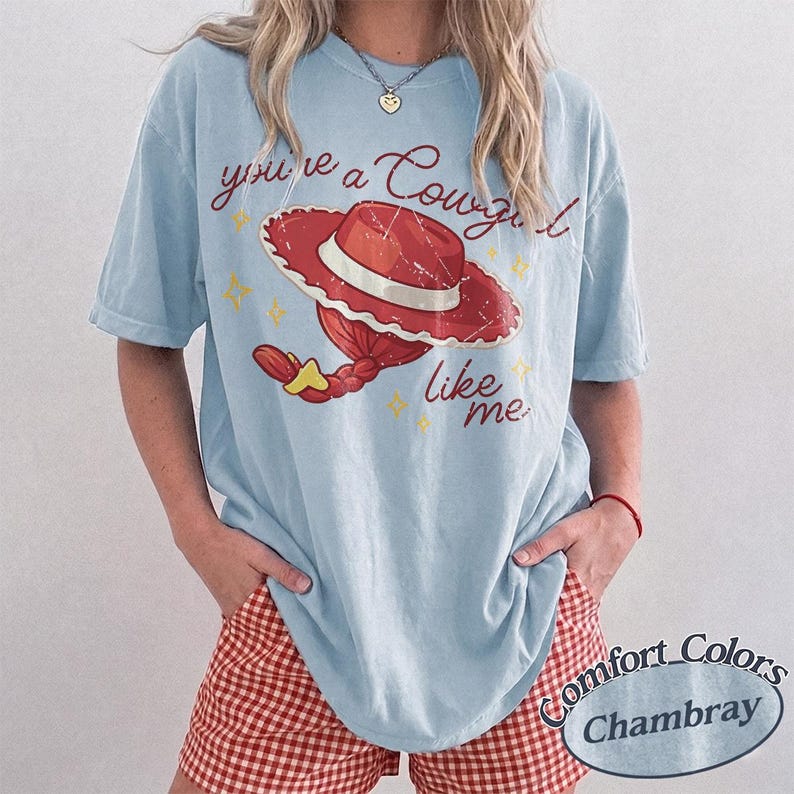May include: A light blue t-shirt featuring a graphic of a red cowgirl hat with a white band and a red braided ponytail. The text reads you're a Cowgirl like me in a cursive red font, accented by small yellow stars. The shirt has a distressed print.